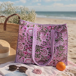 Könnte beinhalten: Eine gesteppte Tragetasche mit einem lebendigen rosa-weißen Blumen-Blockdruckmuster und grünen Akzenten. Die Tasche hat zwei passende Stoffgriffe mit gestreifter Verzierung und eine rechteckige Form mit detaillierten Steppnähten im Stoff.