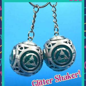 May include: Two spherical keychains featuring a metallic silver cage design with intricate cutouts. Inside each sphere is a translucent green core filled with glitter. The text Glitter Shaker! is printed in yellow with a pink outline at the bottom.