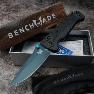 May include: A folding pocket knife with a blue-grey blade and a black textured handle. The knife features a bright blue thumb stud and the Benchmade logo on the blade. It is presented in a branded wooden box with a black fabric storage pouch.