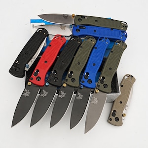 May include: A collection of folding pocket knives with textured handles in red, black, olive green, blue, and tan. Several knives are open, displaying black or silver blades with a butterfly logo, while others are closed and stacked on a box.
