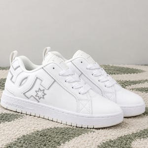 Puede incluir: Un par de zapatillas bajas blancas con un logotipo DC prominente en el lateral, cordones blancos, cuello acolchado y una suela de goma blanca gruesa. El panel lateral muestra un emblema de estrella y la entresuela incluye texto en relieve.