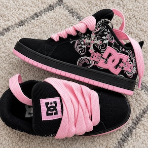 May include: A pair of black and pink DC sneakers featuring thick pink laces, a black suede upper, and a pink rubber sole. The side displays a pink DC logo and a silver cross charm with pink accents. The tongue has a pink patch with a black DC logo.