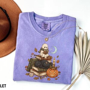 May include: A violet short-sleeved t-shirt with a Comfort Colors label. The graphic features a ghost wearing a scarf reading a book, a black cat sleeping on a stack of books, a pumpkin with a candle, a crescent moon, and scattered autumn leaves.
