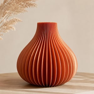 Può includere: Un vaso color terracotta con una distinta texture plissettata a fisarmonica. Il recipiente presenta un corpo arrotondato e bulboso che si restringe verso una stretta apertura circolare in alto, creando un motivo a coste verticali uniforme.