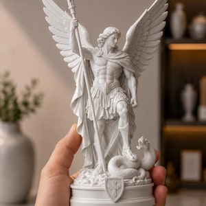 Może przedstawiać: Biała, matowa figurka skrzydlatej postaci w klasycznej zbroi, trzymającej długą włócznię. Postać stoi jedną stopą na zwiniętym wężu, umieszczona na okrągłej podstawie z emblematem tarczy ozdobionym wzorem krzyża, wykonana z detalami.