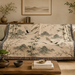 May include: A woven throw blanket featuring a beige background with a repeating pattern of flying cranes, bamboo stalks, and stylized clouds in shades of black, white, and muted green. The blanket has a fringed edge and is draped over a light-colored sofa.