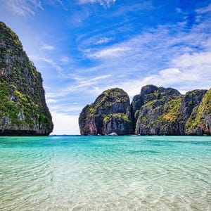 May include: A tropical bay featuring clear turquoise water in the foreground with ripples on the surface. Large, steep limestone cliffs covered in green vegetation rise from the sea under a bright blue sky with scattered white clouds.