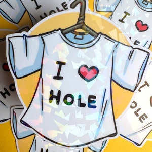Peut inclure: Un t-shirt blanc avec un cœur rouge et le texte "I HOLE" imprimé dessus. Le t-shirt est sur un cintre et a une finition holographique.