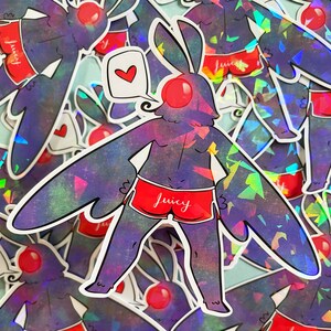 May include: A holographic sticker of a purple creature with bunny ears wearing red underwear with the word "Juicy" printed on it. The creature has a red nose and a speech bubble with a red heart.
