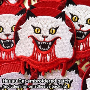 May include: Embroidered patch depicting a white cat with a sinister grin and yellow eyes, framed by red details. The cat has black ears and a red, dripping design. The text "Hausu Cat embroidered patch" is visible.