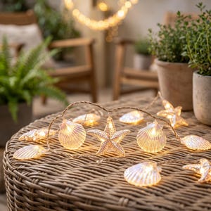 May include: A string of decorative LED lights featuring translucent, iridescent seashell and starfish shapes. The lights emit a warm glow and are connected by a thin, flexible wire, resting on a woven surface with a textured, light brown finish.