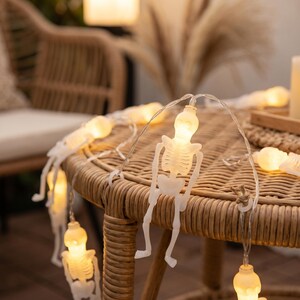 May include: A string of decorative LED lights featuring small, white, translucent skeleton figures. Each skeleton is illuminated from within, emitting a warm, soft glow. The figures are connected by a thin, clear wire, creating a festive lighting display.
