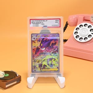 May include: A graded 2025 Pokemon M2a JP Mega Gengar ex trading card encased in a clear plastic slab. The card features a purple Gengar illustration with 350 HP text. The slab is displayed on a white stand, positioned near a pink rotary telephone.