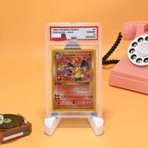Könnte beinhalten: Eine französische Pokemon Charizard Sammelkarte von 1999, eingeschlossen in einer transparenten PSA-Schutzhülle. Die Karte zeigt eine holografische Illustration der orangefarbenen Kreatur. Das Etikett lautet 1999 POKEMON FRENCH DRACAUFEU - HOLO GEM MT.