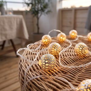 May include: A string of decorative lights featuring spherical, metallic gold-toned covers with a perforated, lattice-like pattern. Each globe emits a warm, soft glow from within, connected by a clear, flexible electrical cord resting on a surface.