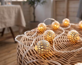 May include: A string of decorative lights featuring spherical, metallic gold-toned covers with a perforated, lattice-like pattern. Each globe emits a warm, soft glow from within, connected by a clear, flexible electrical cord resting on a surface.