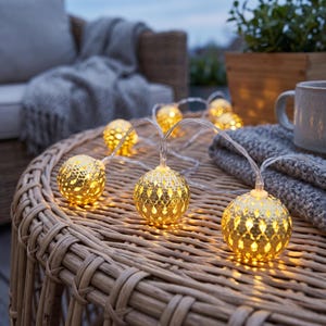 May include: A string of decorative lights featuring spherical gold-toned metal shades with intricate cut-out patterns. Each globe emits a warm, glowing light, connected by a thin, clear electrical cord, creating a festive and ambient illumination.