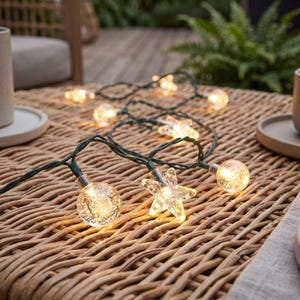 Puede incluir: Una guirnalda de luces decorativas con esferas de vidrio transparente con texturas de burbujas internas y bombillas en forma de estrella. Las luces están conectadas por un cable verde oscuro y emiten un brillo cálido sobre una superficie trenzada.