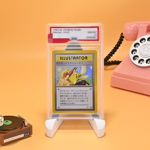 May include: A graded 1998 Japanese promo Pikachu Illustrator trading card encased in a clear plastic slab. The card features an illustration of Pikachu holding a paintbrush and pen, with the text "ILLUSTRATOR" printed at the top in bold letters.