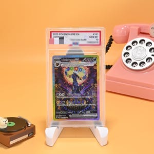 May include: A graded Umbreon GX trading card in a clear plastic slab, featuring a dark creature with a colourful circular aura. The card label reads 2025 POKEMON PRE EN #161 UMBREON GEM MT 10. The card is displayed on a white stand next to a pink rotary phone.