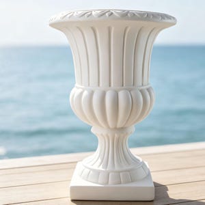 May include: A matte white urn planter featuring a wide flared rim with a decorative scalloped edge, a fluted upper body, a rounded midsection with vertical ridges, and a tapered pedestal base resting on a square plinth for stability.