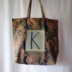 Puede incluir: Un bolso tote con un patrón de tapiz floral oscuro en tonos marrón, naranja y verde. Un parche rectangular central muestra una letra K mayúscula grande de color negro sobre un fondo beige claro, bordeado por un ribete decorativo fino.