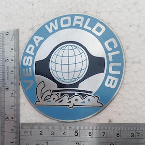 May include: A circular metal emblem featuring the text "VESPA WORLD CLUB" in silver on a light blue background. A central globe graphic sits above the Vespa logo. A stainless steel ruler shows the emblem has a diameter of approximately 9 cm.