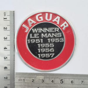 May include: A circular metal emblem featuring a red outer ring with the word JAGUAR in silver letters. The black center displays the text WINNER LE MANS 1951 1953 1955 1956 1957. A metal ruler shows the emblem has a diameter of approximately 7.5 cm.
