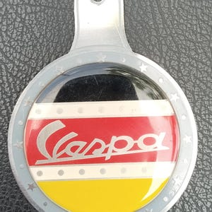 May include: A circular metal emblem featuring a glossy center with horizontal black, red, and yellow stripes. The word Vespa is written in silver script across the center. The outer silver border is decorated with small embossed stars and dots.