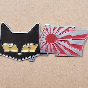Puede incluir: Un pin de esmalte metálico que presenta una cabeza de gato negra estilizada con ojos amarillos en el lado izquierdo, conectada a una forma rectangular a la derecha que muestra un sol rojo con rayos rojos y plateados sobre fondo plateado.