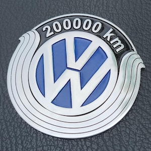 Könnte beinhalten: Ein kreisförmiges Emblem aus Metall mit dem Volkswagen-Logo in Blau und Silber. Das Design umfasst einen schwarzen Bogen am oberen Rand mit dem silber geprägten Text 200000 km. Das Emblem ist von stilisierten silbernen Wellenmustern umgeben.