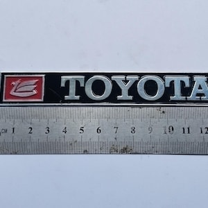 May include: A rectangular Toyota emblem featuring silver lettering on a black background. On the left side, a red square displays a silver stylized wing logo. A metal ruler positioned below indicates the emblem is approximately 14 cm in length.