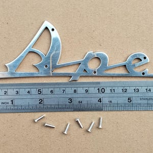 May include: A silver-colored metal script emblem reading "Alpine" with a brushed finish, featuring several small mounting holes. It is positioned next to a stainless steel ruler showing a width of approximately 6.5 inches. Six small metal rivets are included.