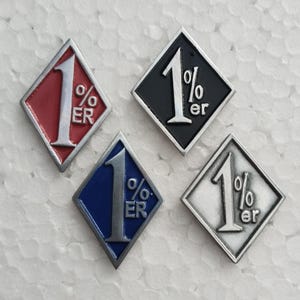 Pode incluir: Quatro pins de metal em forma de diamante com o texto 1%er em relevo prateado. Os pins têm fundos de esmalte colorido em vermelho, preto, azul e prata, cada um emoldurado por uma borda prateada com um design pontiagudo no topo e na base.