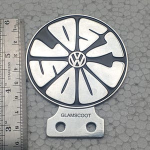 Könnte beinhalten: Ein kreisförmiges Emblem aus silbernem und schwarzem Metall mit einem stilisierten Volkswagen-Logo in der Mitte und dem Wort "LOST" im Design. Eine Metallhalterung mit zwei Löchern und der Aufschrift "GLAMSCOOT" ist angebracht. Ein Lineal zeigt einen Durchmesser von etwa 9 cm.