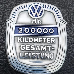 Könnte beinhalten: Ein silber-blaues Volkswagen-Metallabzeichen mit dem VW-Logo oben. Der Text lautet FÜR 200000 KILOMETER GESAMT-LEISTUNG. Das schildförmige Emblem hat eine gebürstete Metalloberfläche mit erhabener blauer Schrift und einem blauen Rand.
