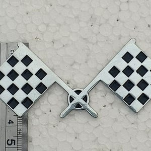 May include: A silver-toned metal emblem featuring two crossed checkered flags with black squares. A metal ruler is positioned along the left side, showing the emblem measures approximately 2.5 inches in width and 1.5 inches in height.