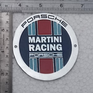 Könnte beinhalten: Ein kreisförmiges Porsche Martini Racing Emblem aus Metall mit dunkelblauem Hintergrund und vertikalen roten sowie hellblauen Streifen. Der Text PORSCHE, MARTINI RACING und PORSCHE ist in Weiß gedruckt. Ein Metalllineal zeigt 8,9 cm Durchmesser.