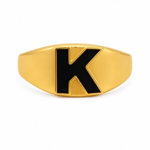 以下が含まれることがあります： 中央に目立つ太字の黒い文字「K」が配置された、平らな楕円形の面を持つゴールドトーンのシグネットリング。リング部分は滑らかで磨き上げられており、暗いインレイと対照的な反射性のある金属仕上げが施されています。
