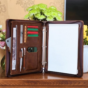May include: A brown leather-look zipped portfolio organiser featuring a three-ring binder mechanism, a grid-lined notepad, multiple card slots, two pen loops, a zipped document pocket, and dedicated compartments for a smartphone and tablet.