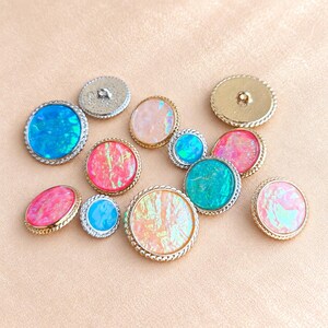 May include: A collection of round buttons featuring iridescent, opal-like inserts in shades of blue, pink, and peach. Each button is framed by a metallic, twisted rope-style border. The buttons vary in size and display a smooth, reflective surface.