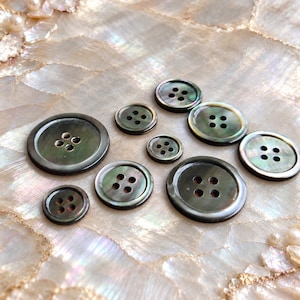 May include: A collection of nine round, dark-toned mother-of-pearl buttons of varying sizes, each featuring a smooth, polished surface with a subtle iridescent sheen and four centered holes for attachment, arranged in a scattered formation.