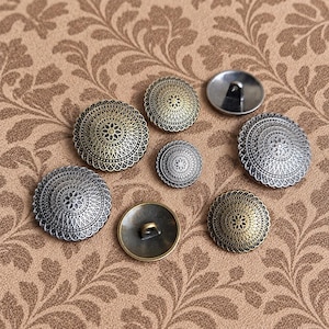 May include: A collection of seven round metal buttons featuring intricate geometric patterns and floral centers. The buttons are finished in varying shades of antique gold and silver, with some showing the smooth back and loop shank attachment.