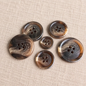 Peut inclure: Un ensemble de cinq boutons ronds de tailles variées, présentant chacun un motif marbré brun foncé et beige. Chaque bouton possède quatre trous au centre et un rebord extérieur surélevé, avec une finition lisse et polie sur l'ensemble.
