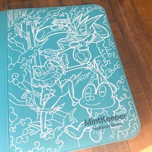 May include: A teal-coloured MintKeeper toploader binder featuring a white line-art illustration of three frog-like creatures amidst stylised clouds and water elements. The cover includes the text MintKeeper Toploader Binder in white lettering.
