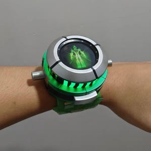 May include: A chunky green and silver toy watch featuring a circular face with a glowing green digital display showing a humanoid figure. The device has a thick, segmented green band and metallic silver accents around the central dial mechanism.