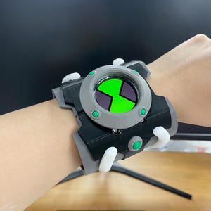 May include: A chunky wrist-worn device featuring a circular grey frame with a bright green and black hourglass-shaped display. The accessory includes black and grey structural components, accented by small, protruding white and green circular buttons.