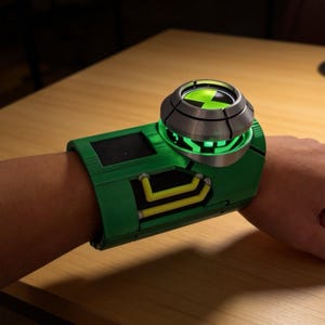 Puede incluir: Un dispositivo verde para la muñeca con un dial circular plateado y un emblema central verde brillante. El brazalete incluye una pantalla rectangular negra y líneas amarillas, con una luz verde intensa que emana bajo el dial central.