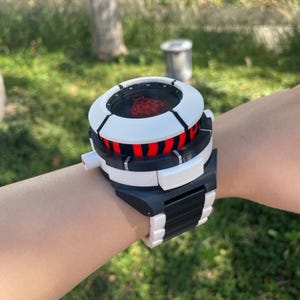 May include: A chunky, oversized wristwatch featuring a thick white and black frame with a circular face. The display shows a red graphic of a stylized figure. The watch band consists of alternating black and white segments, creating a bold, mechanical appearance.