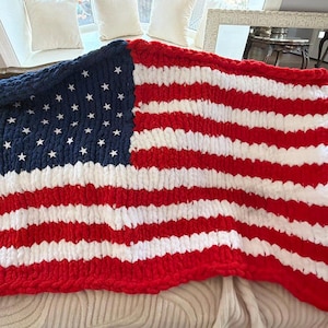 May include: A chunky knit blanket featuring the American flag design. The blanket is made of thick, soft yarn in red, white, and blue. The blue section in the upper left corner contains white star shapes, while the rest of the blanket displays horizontal red and white stripes.